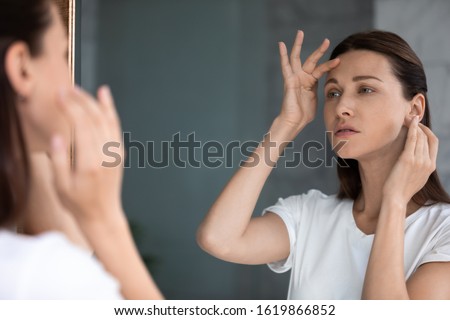 Anxious woman looking in mirror, standing in bathroom, upset attractive female touching forehead with finger, confused about wrinkles or acne, thinking about face lifting procedure or spa Royalty-Free Stock Photo #1619866852