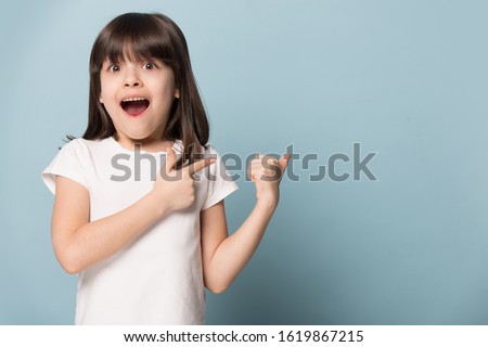 Excited gawp six years old little brown-haired girl pointing back on empty copy space for advertisement text of new toys store, children goods, services or events, isolated on blue studio background. Royalty-Free Stock Photo #1619867215