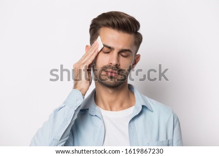 Isolated on white studio background overheated sweaty young man suffering from heat stroke or high temperature, using paper napkin. Head shot close up portrait tired millennial guy feeling unhealthy. Royalty-Free Stock Photo #1619867230