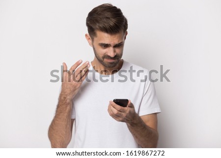 Isolated on white studio background disappointed millennial guy holding smartphone, annoyed by trouble with wireless connection, slow internet service, broken mobile gadget, received bad news email.