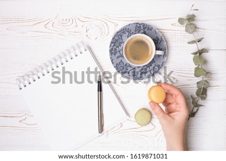 Female hand holds macaroon, a cup of coffee and a clean diary with spring and a branch of eucalyptus on a white shabby wooden table. Fashion flat lay.