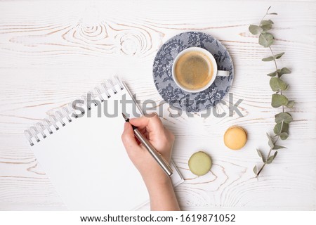 A female hand holds a pen over a clean diary with spring, a cup of coffee and macaroons and a branch of eucalyptus on a white shabby wooden table. Fashion flat lay.