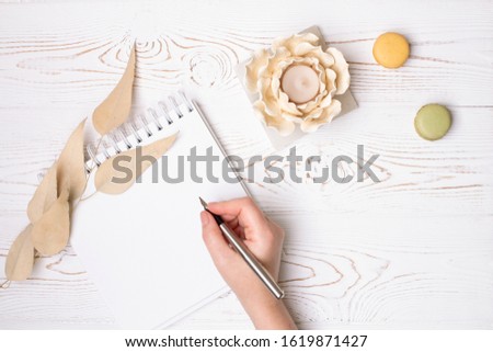 A female hand holds a pen over a clean diary with spring, macaroons, beige leaves and a flower-shaped candlestick on a white shabby wooden table. Fashion flat lay.