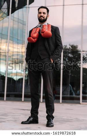 Vertical photo of a businessman in a suit with his hands in his trouser pockets and red boxing gloves hanging from his neck with a building in the background