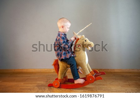 Funny boy in plaid shirt with toy sword and shield made of straw sits on rocking horse. Boy dreams of battles, victories and adventures. Concept of education morale, patriotism, strength of mind