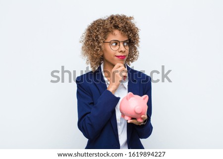 young african american woman smiling with a happy, confident expression with hand on chin, wondering and looking to the side with a piggy bank