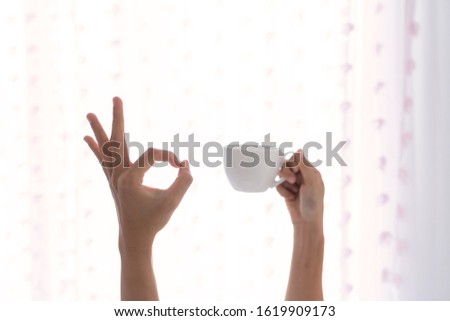 Young beautiful  woman waking up in the morning in the bed, stretching out one's arms with a cup of coffee and showing OK sign.