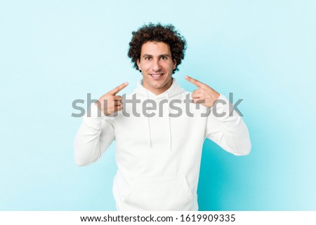 Young sporty man against a blue background smiles, pointing fingers at mouth.