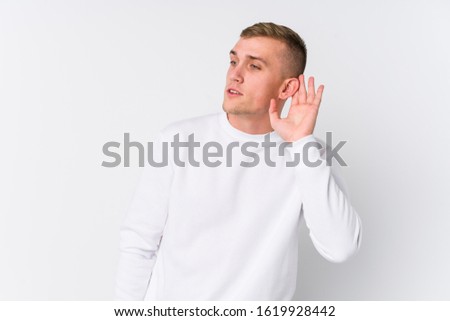 Young caucasian man on white background trying to listening a gossip.
