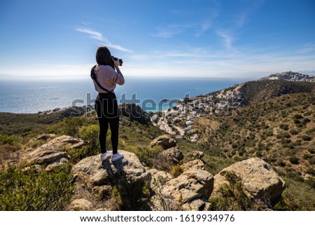 Beautiful woman photographer standing on a cliff taking a picture with a professional camera with the mediterranean sea in the background in Spain with copy space