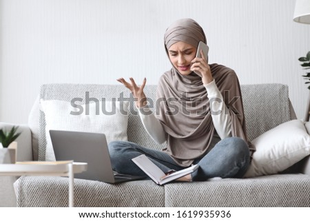 Freelance stress. Concerned muslim girl in hijab talking on cellphone and emotionally gesturing while working online on laptop at home, free space