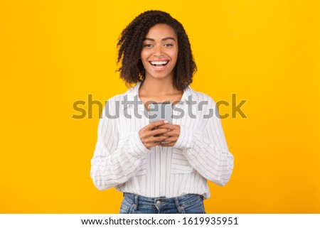 Chatting With Friends. Happy black woman holding mobile phone, looking at camera over yellow background, free space