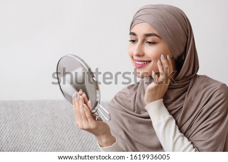 Beauty routine. Attractive muslim girl in hijab holding mirror and looking at her face with perfect soft skin, sitting on sofa at home, free space Royalty-Free Stock Photo #1619936005