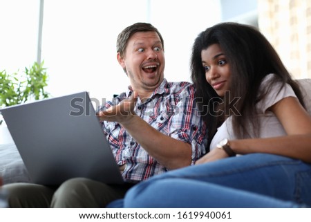 Portrait of surprised man showing something to african-american woman on laptop. Astonished female sitting next to husband on couch. Mixed race family concept
