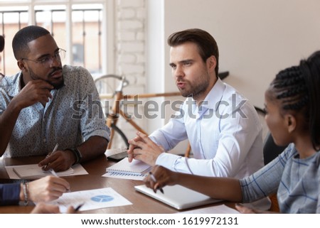 Serious Caucasian man worker talk discussing ideas with diverse colleagues at team meeting in office, millennial multiracial businesspeople coworkers brainstorm at briefing, cooperation concept Royalty-Free Stock Photo #1619972131