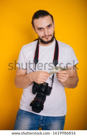 The photographer is holding money. A guy in a white T-shirt with a camera on a yellow background holds dollars in his hands. Photographer Salary