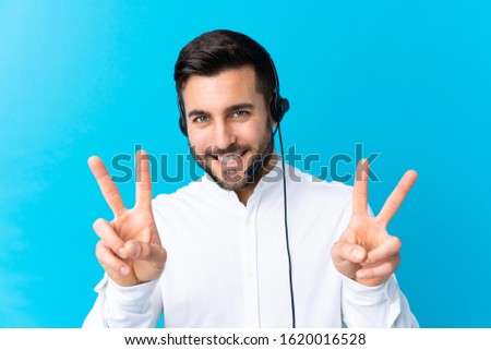 Telemarketer man working with a headset over isolated blue background smiling and showing victory sign