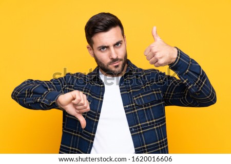 Young handsome man with beard over isolated yellow background making good-bad sign. Undecided between yes or not