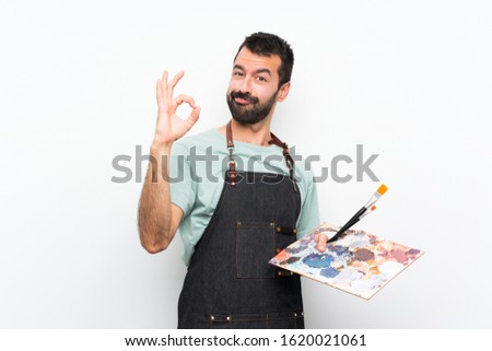 Young artist man holding a palette over isolated background showing ok sign with fingers