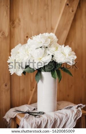 Stylish peony bouquet in white ceramic vase on linen fabric with scissors on rustic wooden background.White peonies rural still life. Hello spring wallpaper. Happy Mothers day. Space