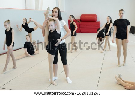 Young female coach assisting flexible girl to do split standing on one leg while other girls performing warm-up exercises
