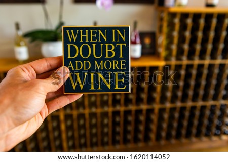 When in doubt add more wine, hand holding a quote sign selective focus, tasting room shop interior, wines bottles on wooden shelves rack background