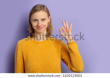 portrait of young caucasian woman showing OK gesture at camera, wearing yellow casual blouse and smile, isolated over purple background
