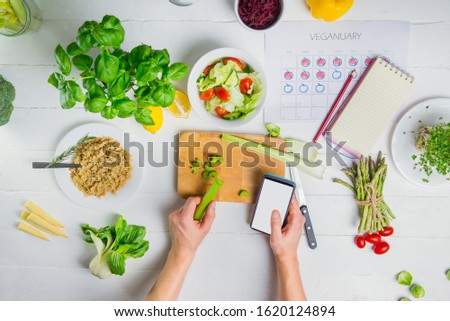 Top view Man's hands holding smartphone with blank white screen and celery stick above the cutting board. Cooking vegan fresh salad with vegetables. Veganuary calendar and daily diet planning