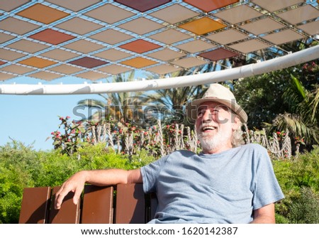 A senior people with white beard and hair smiling and relaxing sitting on a bench. Tropical plants around him. Blue sky