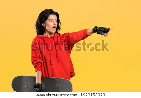 Young skater woman surprised and pointing finger to the side over isolated background