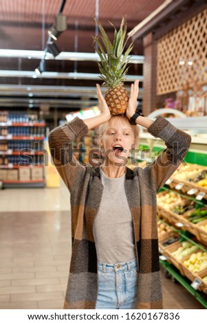 Young woman at the supermarket doing daily shoppings standing holding pineapple above head singing having fun silly Royalty-Free Stock Photo #1620167836