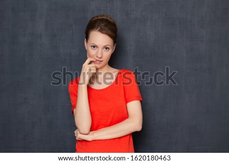 Studio portrait of focused thoughtful young blond woman wearing T-shirt, holding hand near lips, looking tiredly at interlocutor, listening or waiting something, standing against gray background