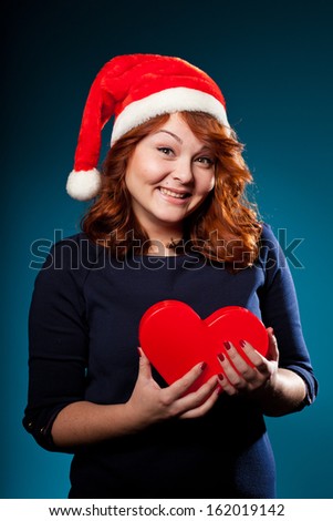 Young lovely smiling red-haired woman in Santa's hat keeping a red heart