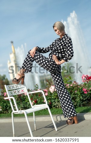 tall beautiful blonde European woman in a black trouser suit with white spots, pants and a shirt, high heel. Short haircut, bright makeup, long legs. lessons Stretching, twine outdoors
