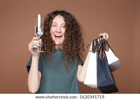 Young excited female with festive hairstyle, feels relaxed, rising glass with cold non alcoholic drink or water, looks happily, holding shopping bags. People, lifestyle, leisure and  holidays concept.