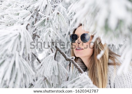 close-up young beautiful caucasian model with long hair in a gray knitted sweater and sunglasses hid in the branches of a snowy pine
