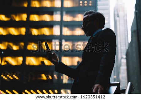 From below side view of middle aged businessman in business suit standing in shadow of skyscrapers and holding smartphone in hand in New York Royalty-Free Stock Photo #1620351412