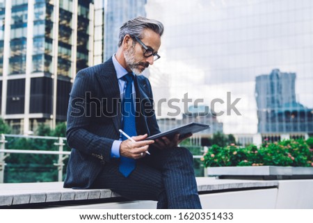 Side view of handsome concentrated middle aged man in glasses and business suit using tablet with stylus while sitting against background of New York structures