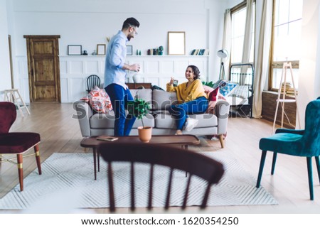 Side view of tall young man with cup of coffee in hand standing next to sofa and talking to laughing woman sitting on couch
