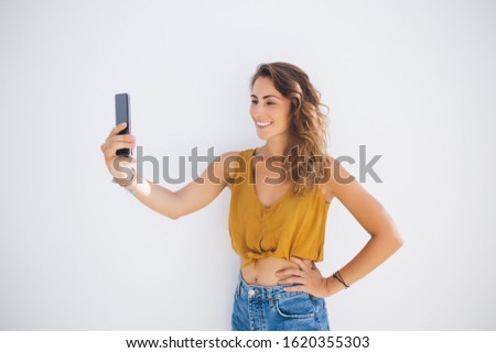 Smiling stylish beautiful adult female in modern summer wear capturing selfie picture by mobile while putting hand on waist on white background 