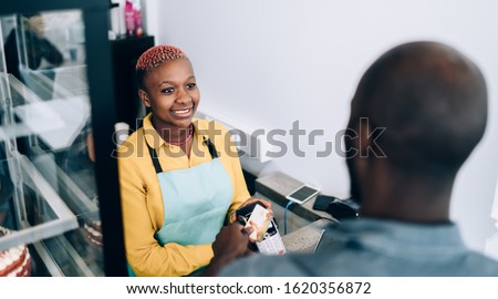 From above smiling African American seller holding terminal and waiting while client making contactless payment with credit card standing beside counter in modern pastry shop