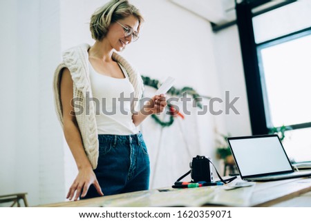 Self employed hipster girl in optical spectacles for vision correction looking at photo image made with instant camera and smiling standing on own studio near desktop with blank laptop computer