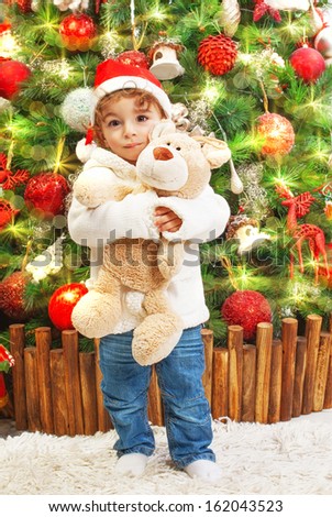 Picture of happy small boy holding teddy bear in hands on beautiful decorated Christmas tree background, little child wearing red Santa Claus hat, New Year celebration, child's xmas party