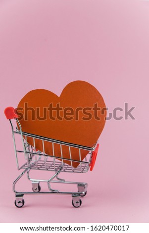 Red heart in a shopping cart on trendy pink background with copy space for text. Concept of holiday Valentine`s day shopping, sale and waste of money for love 