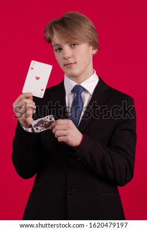 Teenager boy in a black jacket holds playing cards for poker and tricks in his hands and poses on a red background