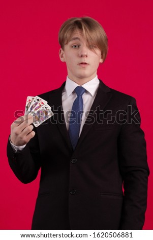 Teenager boy in a black jacket holds playing cards for poker and tricks in his hands and poses on a red background
