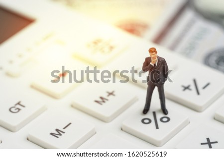 Miniature people business man standing and thinking on calculator percentage button with background of US Dollar banknote using as FED consider interest rate, business profit or stock market analyst.