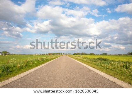 Road in Holland countryside, perspective, under cloudy skies and between green meadows and a faraway straight horizon. Royalty-Free Stock Photo #1620538345