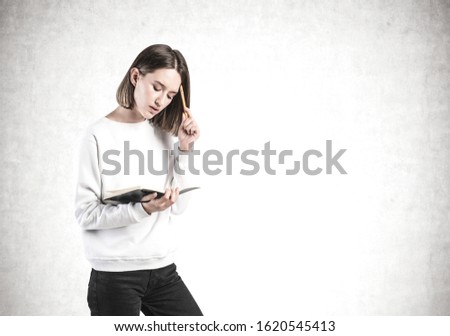Portrait of thoughtful young girl in casual clothes with notebook and pen standing near cocnrete wall. Concept of education and brainstorming. Mock up