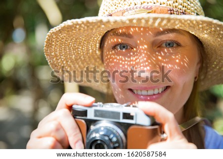 Close-up portrait of a beautiful woman in straw hat travelig in tropic forest, taking photos on retro camera. Light shadows through the cut-out detailed brim on face. Tourist with backpack. 